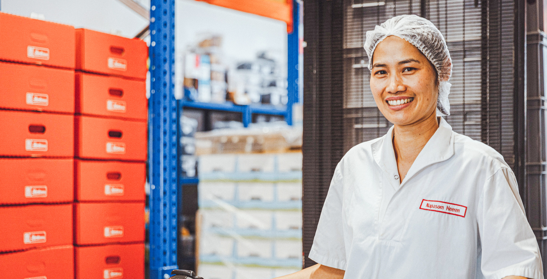 Person trägt weiße Arbeitskleidung mit Haarnetz, steht in einem Lager mit roten Kisten und lächelt in die Kamera