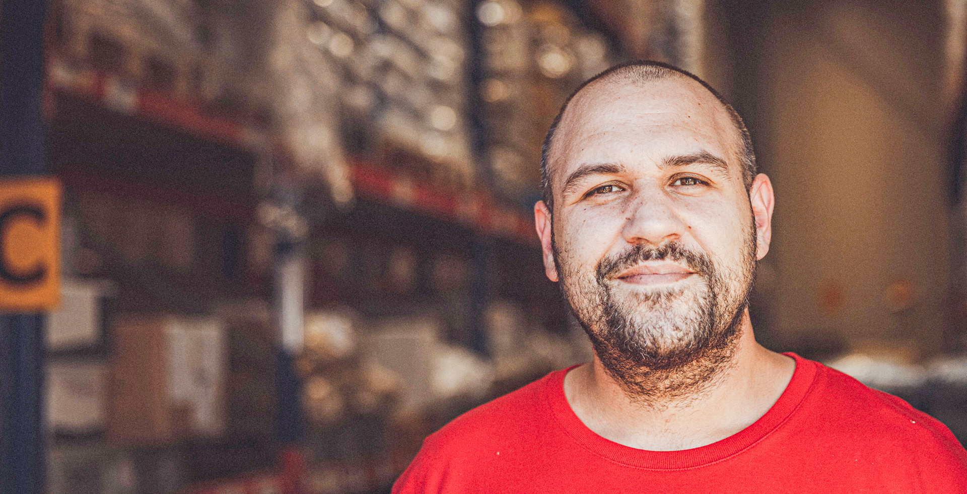 ChatGPT: Mann mit Bart und rotem T-Shirt lächelt in Lagerhalle mit Regalen und Kartons im Hintergrund.