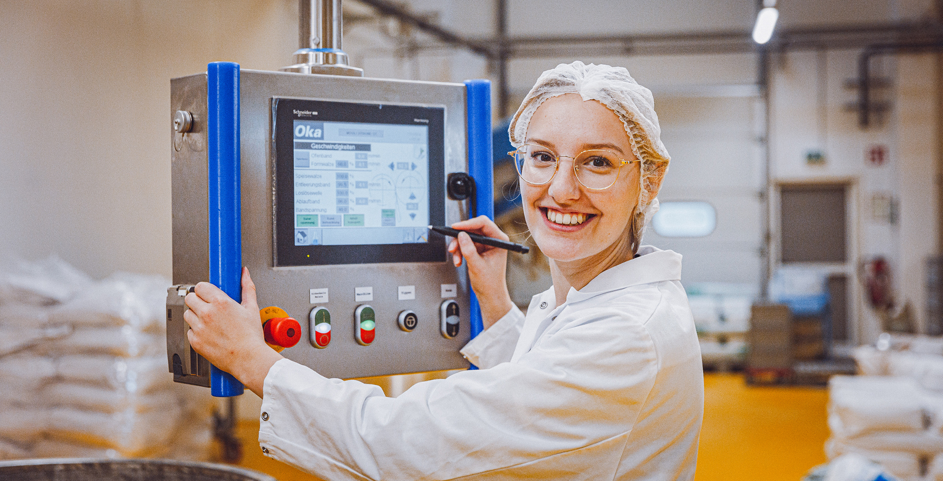 Frau mit Schutzhaube und Brille bedient Kontrollpanel in Produktionshalle und lächelt in die Kamera