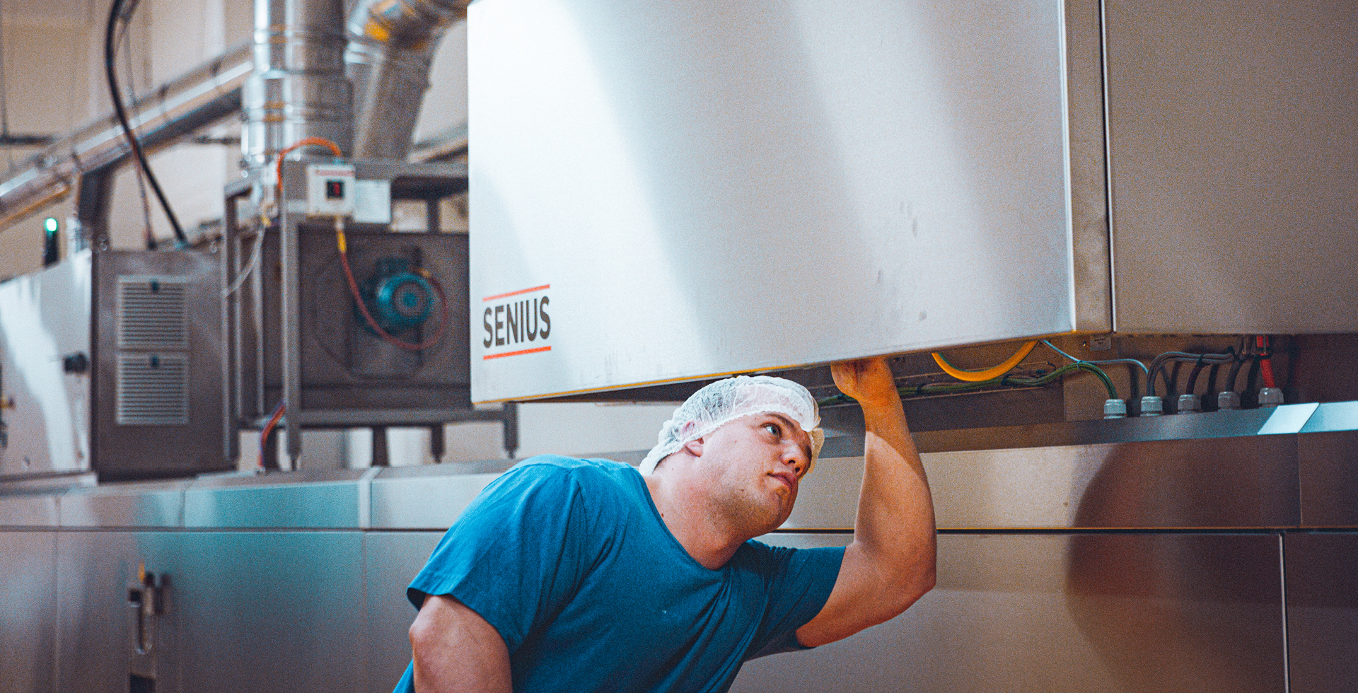 Mann in blauem T-Shirt und Haarnetz inspiziert eine industrielle Maschine mit Aufschrift "SENIUS" in einer Fabrikumgebung