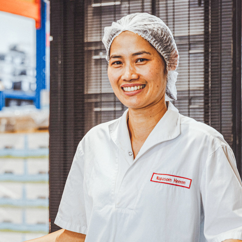 Person trägt weiße Arbeitskleidung mit Haarnetz, steht in einem Lager und lächelt in die Kamera