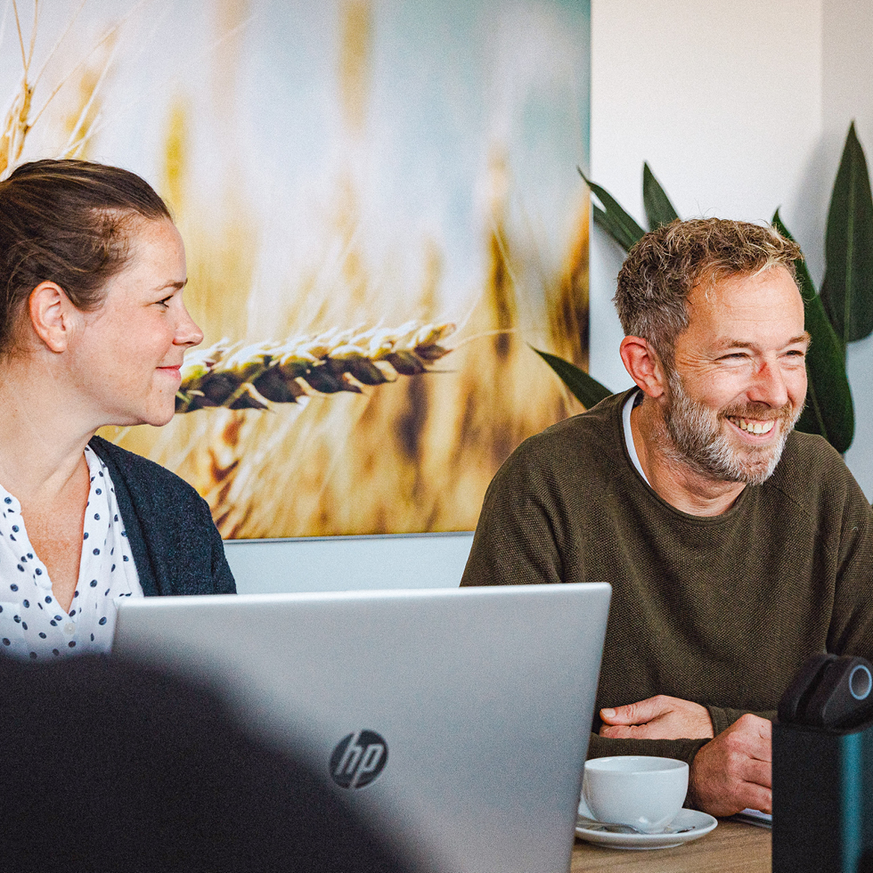 Personen im Meeting, zwei lächeln, Frau mit Laptop, im Hintergrund Bild von Weizen und Pflanze