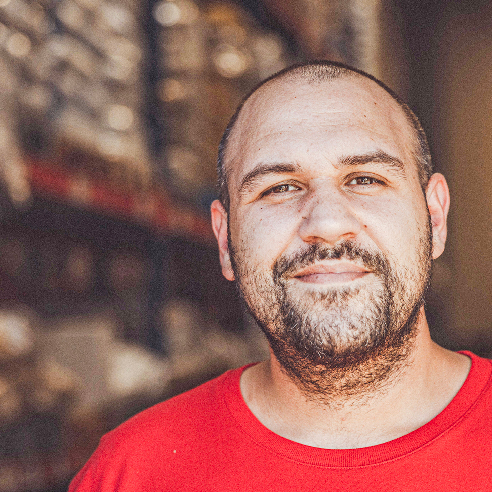 ChatGPT: Mann mit Bart und rotem T-Shirt lächelt in Lagerhalle mit Regalen und Kartons im Hintergrund.