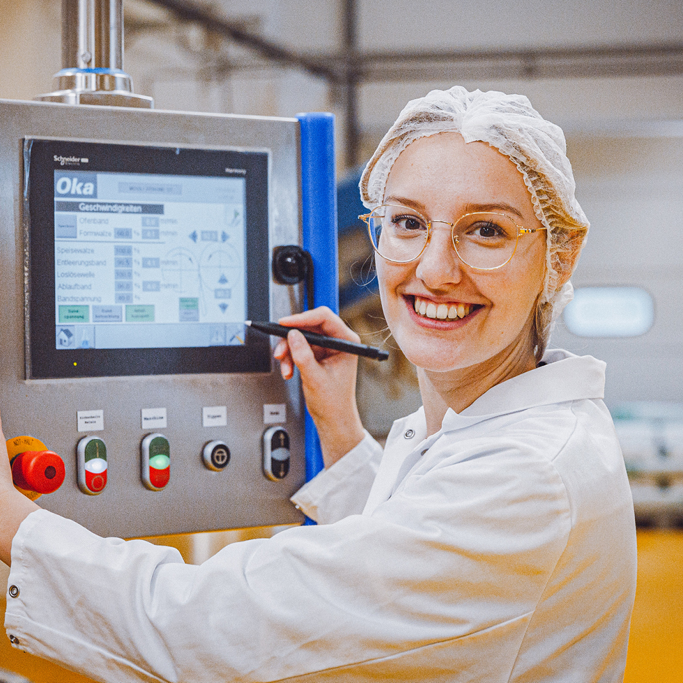 Frau mit Schutzhaube und Brille bedient Kontrollpanel in Produktionshalle und lächelt in die Kamera