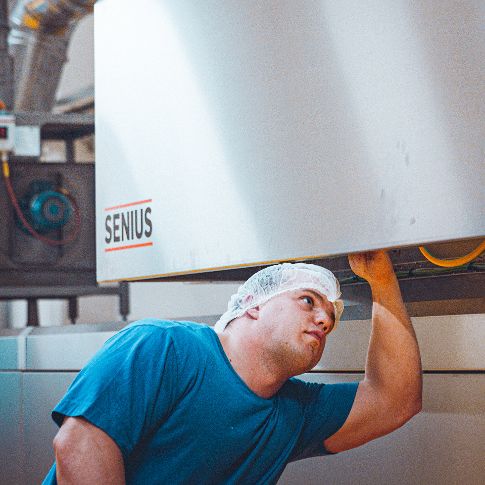 Mann in blauem T-Shirt und Haarnetz inspiziert eine industrielle Maschine mit Aufschrift "SENIUS" in einer Fabrikumgebung