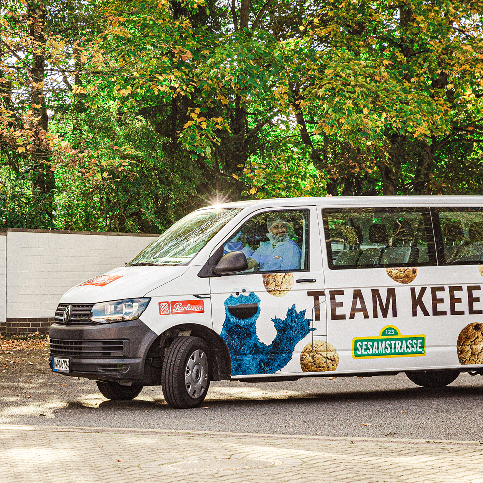 Weißer Van mit Krümelmonster-Motiv und Keksen steht vor Hecke, Fahrer schaut aus Fenster