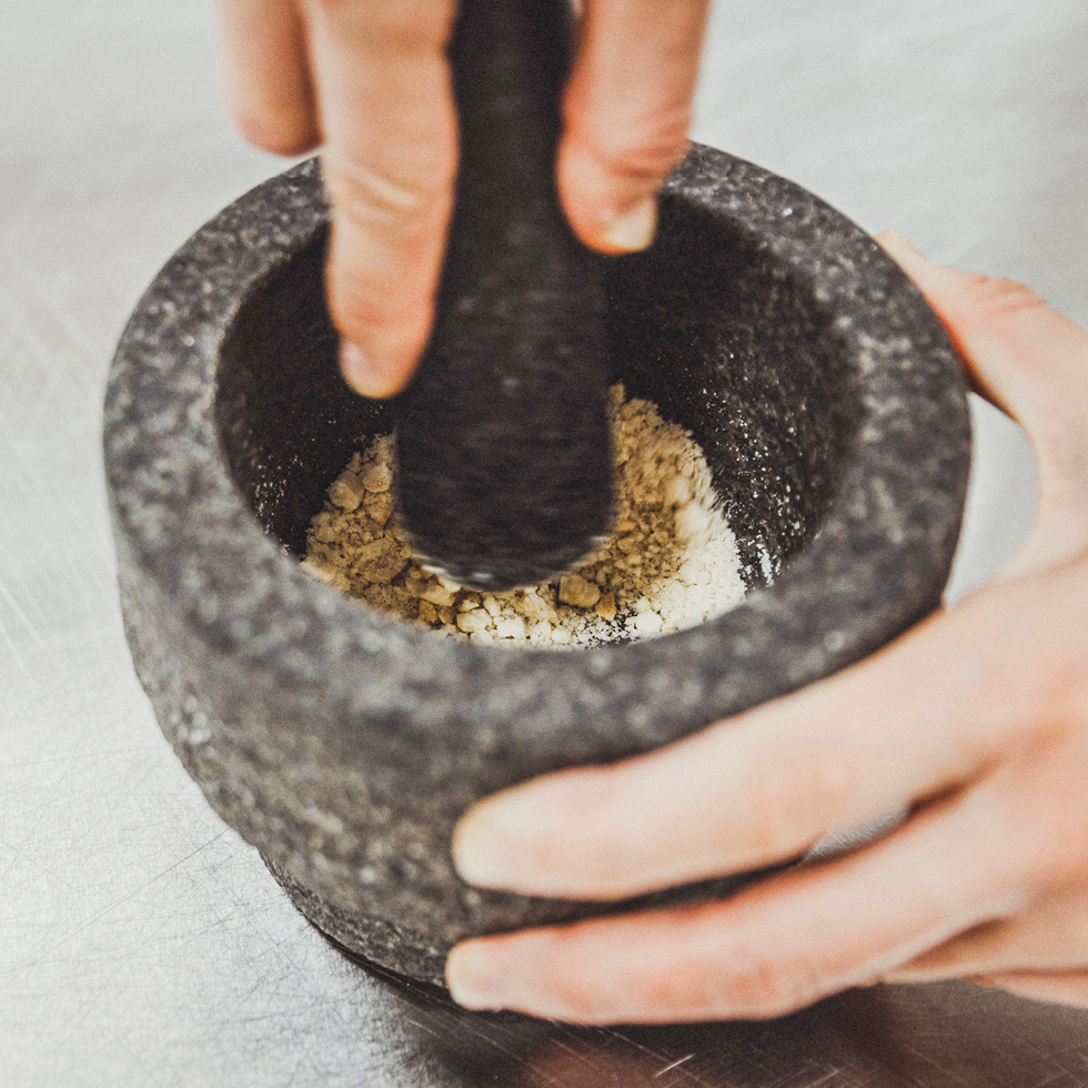 Zwei Hände mahlen Getreide in einem grauen Mörser zu Mehl