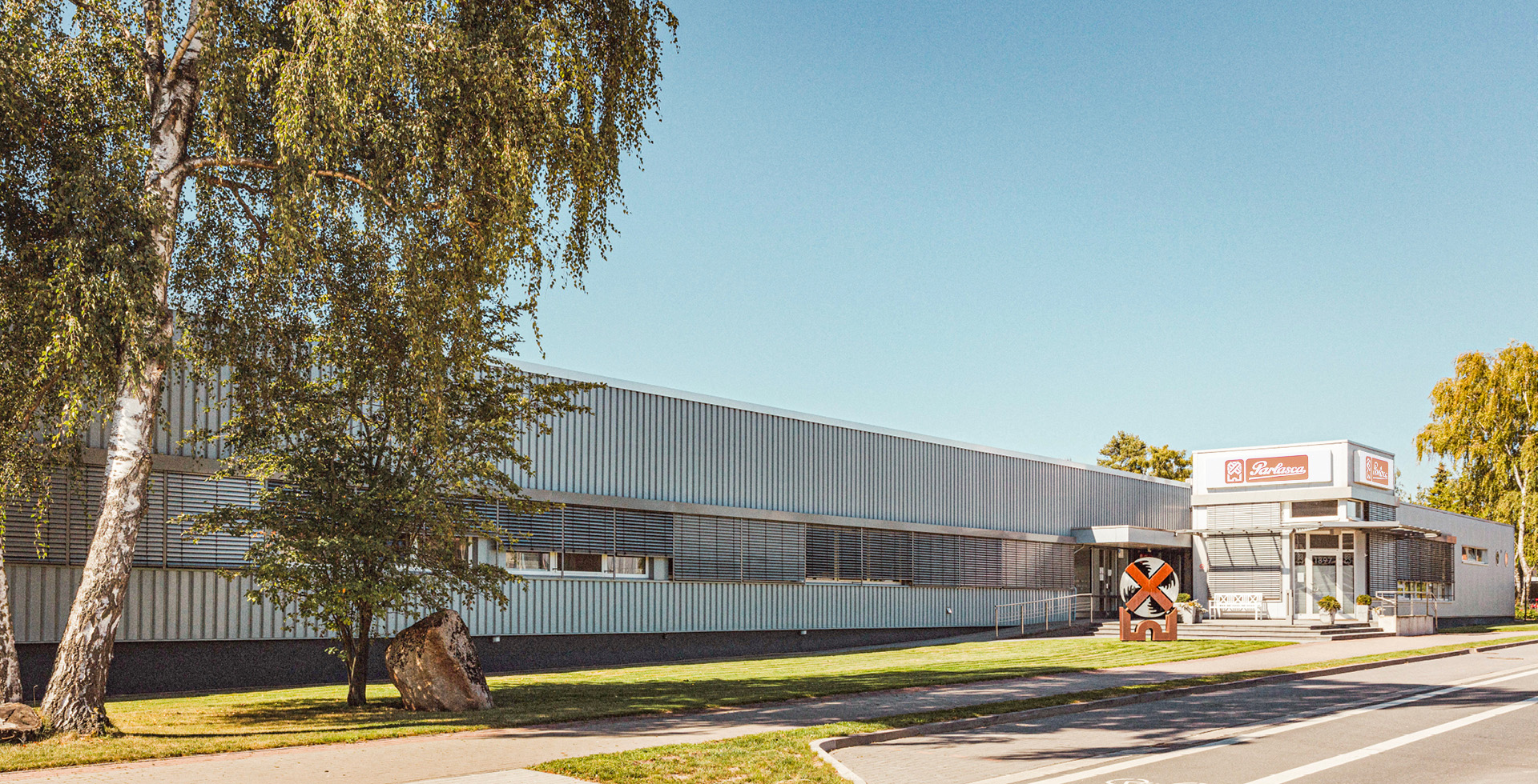 Gebäude der Keksfabrik von außen, große Birke im Vordergrund und Mühlenskulptur auf Rasen