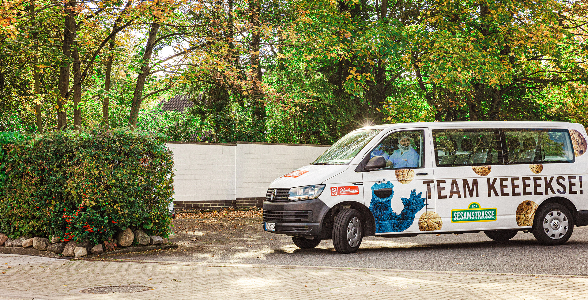 Weißer Van mit Krümelmonster-Motiv und „TEAM KEEEEEKSE!“ steht vor Hecke, Fahrer schaut aus Fenster