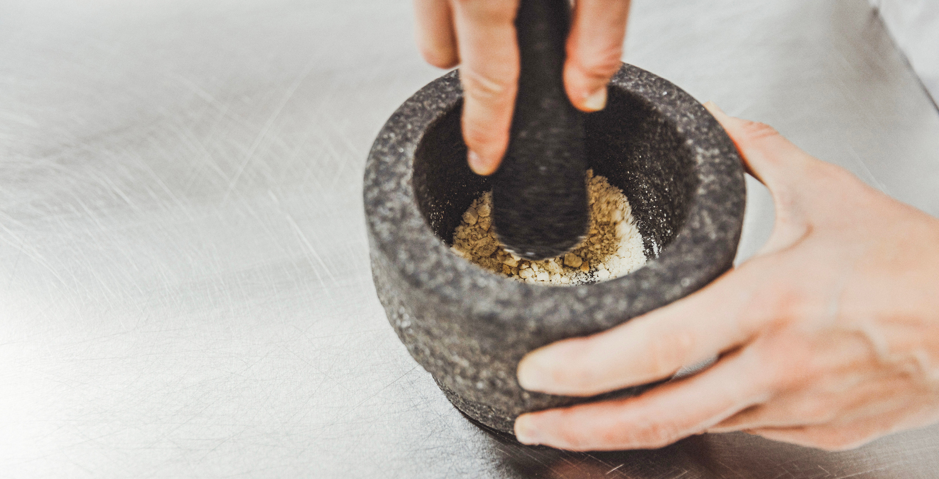 Zwei Hände mahlen Getreide in einem grauen Mörser zu Mehl