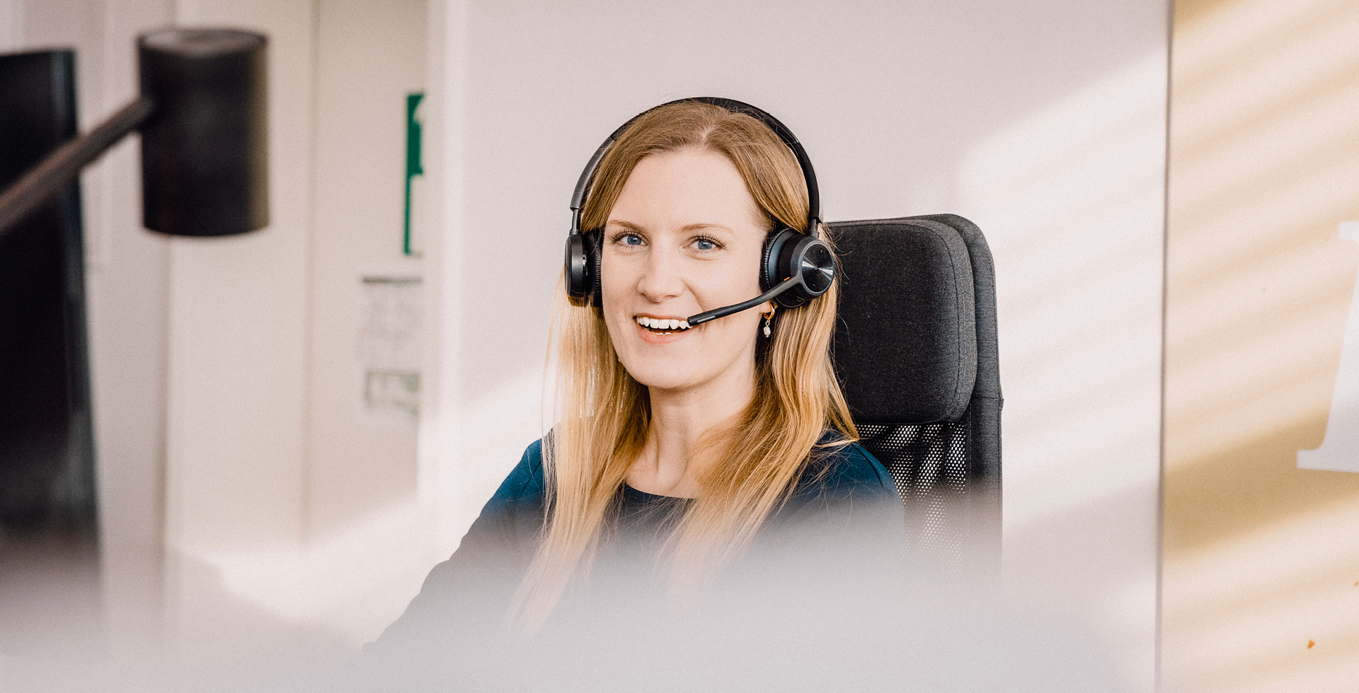 Freundliche Mitarbeiterin mit Headset im Kundenservice, sitzt lächelnd an ihrem Arbeitsplatz im Büro